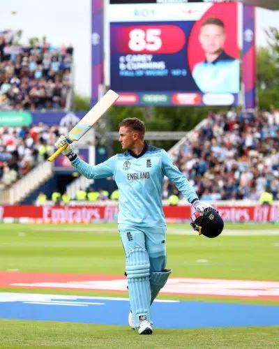 England cricketer in a blue kit raises his bat on the field after scoring, with a cheering crowd and a large scoreboard in the background.