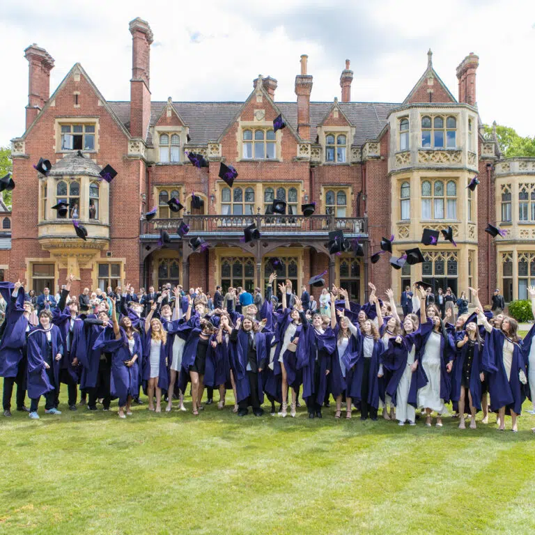 Graduates in purple gowns throwing caps in the air outside a historic brick campus building.