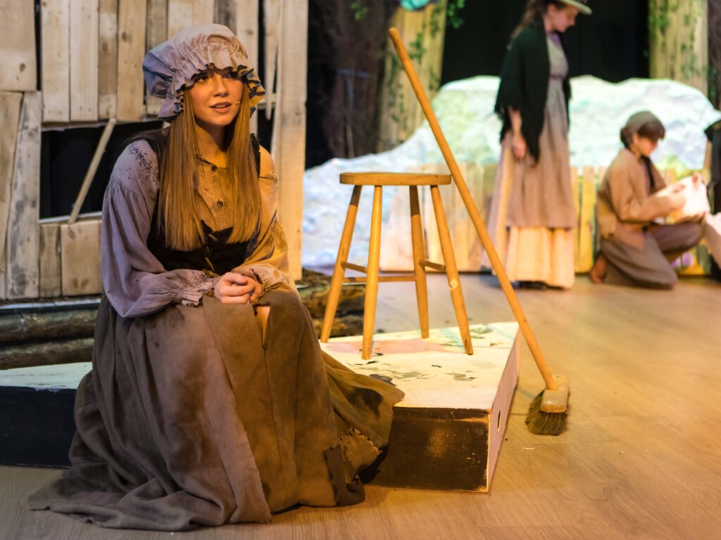 Actor in a period costume with a bonnet sits on a stage platform, hands folded, wooden props nearby.
