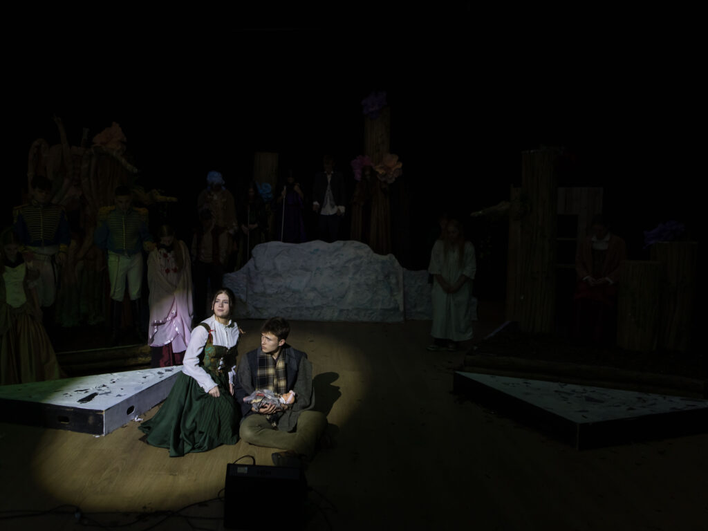 Stage scene with performers in period costumes kneeling in foreground; dark background with props and others standing on set pieces behind a snow-like mound.