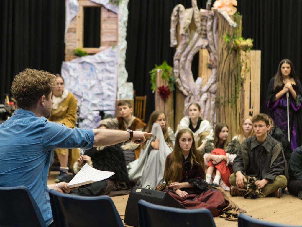 Theatre rehearsal scene: performers in costumes sit on stage with a forest-like set, while a man in a blue shirt at the front points or gestures towards them.