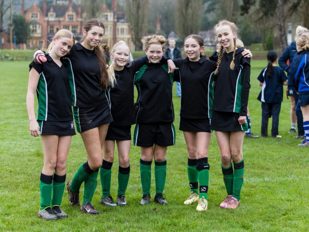 Group of young female players in black and green uniforms posing on a grassy field, smiling at the camera.