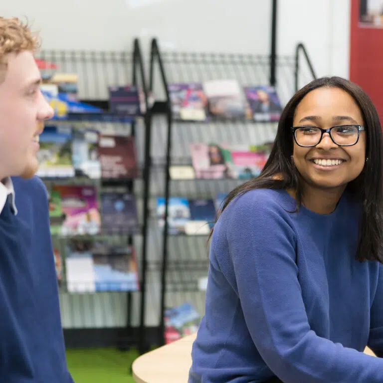 Smiling woman with glasses in a blue sweater sits at a table, man blurred on the left, magazines on shelves behind them.