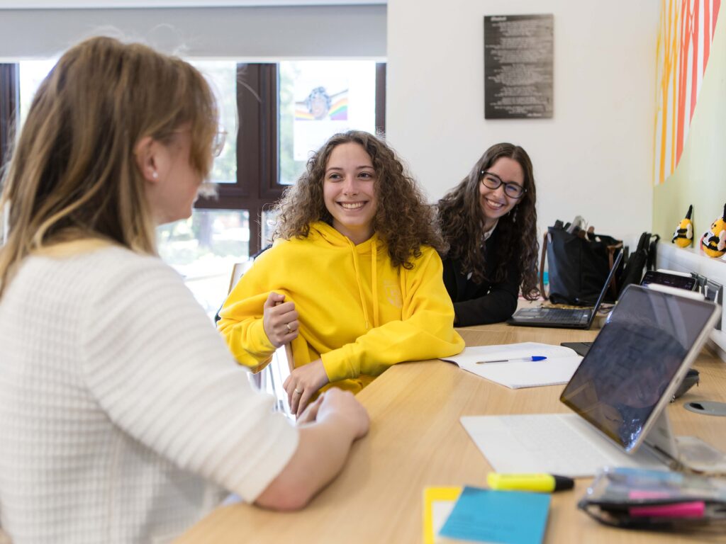 Three students sit at a bright classroom table, smiling and talking with laptops and notebooks open.