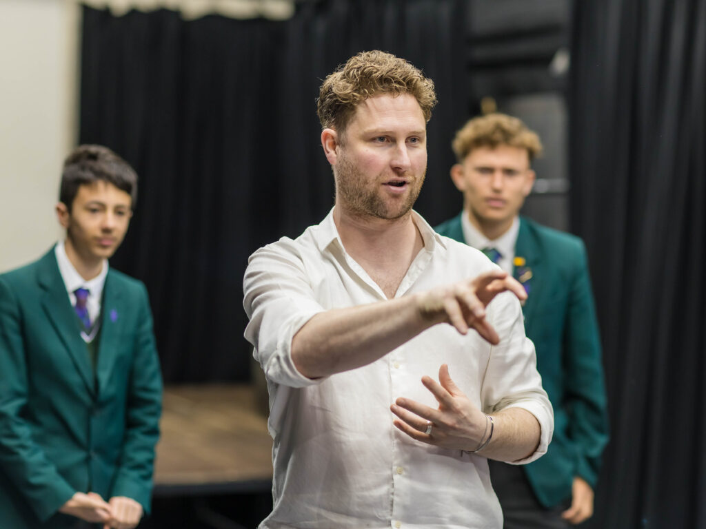 Man in a white shirt gestures with his hands at the camera, while two young men in green blazers stand in the background on a stage.