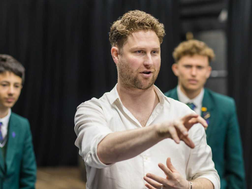 Man in a white shirt gestures with his hands at the camera, while two young men in green blazers stand in the background on a stage.