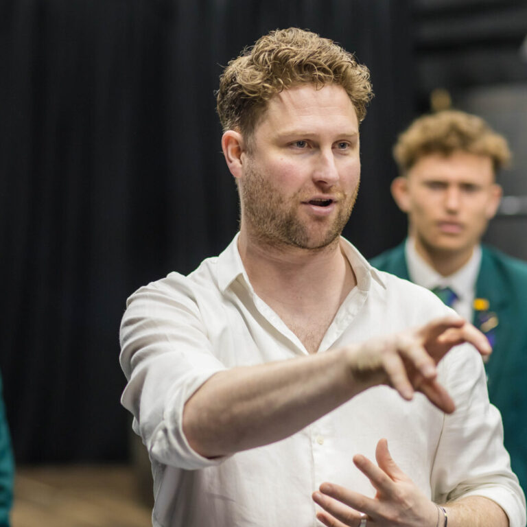 Man in a white shirt gestures with his hands at the camera, while two young men in green blazers stand in the background on a stage.