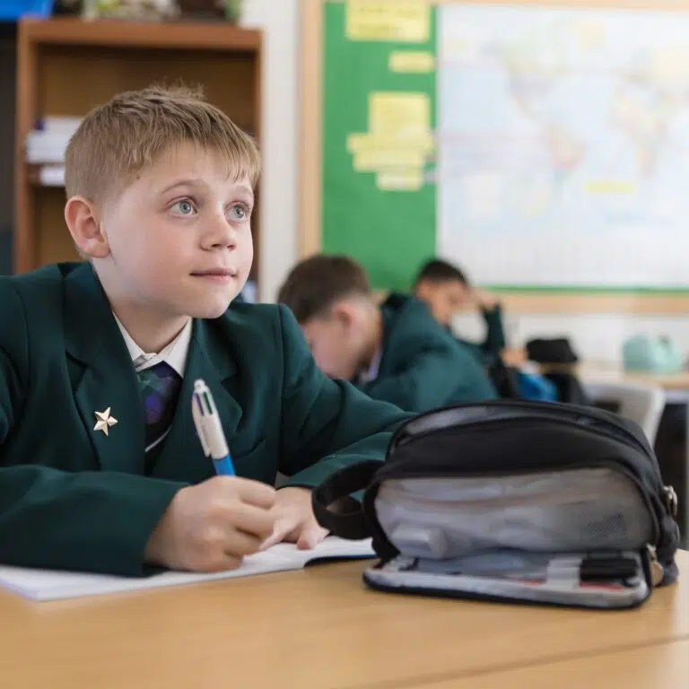 Boy in a green uniform sits at a desk, writing in a notebook in a classroom with a backpack nearby.