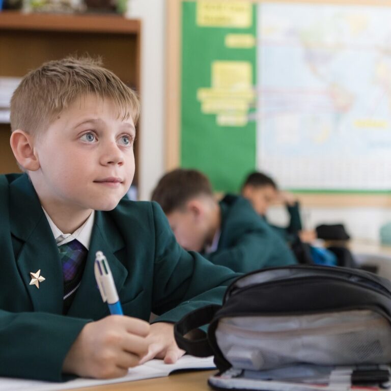 Boy in a green uniform sits at a desk, writing in a notebook in a classroom with a backpack nearby.