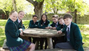 Seven students in green blazers and plaid skirts/shirts sit around a round wooden table in a sunlit park, smiling at the camera.