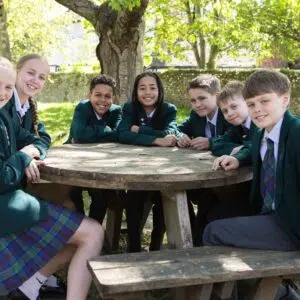 Group of students in green blazers around a wooden picnic table in a sunny park setting.