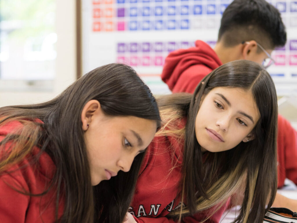 Three students in red hoodies work on worksheets at a bright classroom desk with papers and pencils nearby.