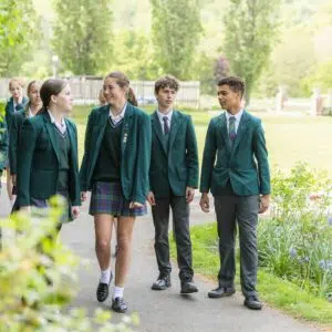 Group of students in green blazers walking and talking along a sunny campus path surrounded by greenery.