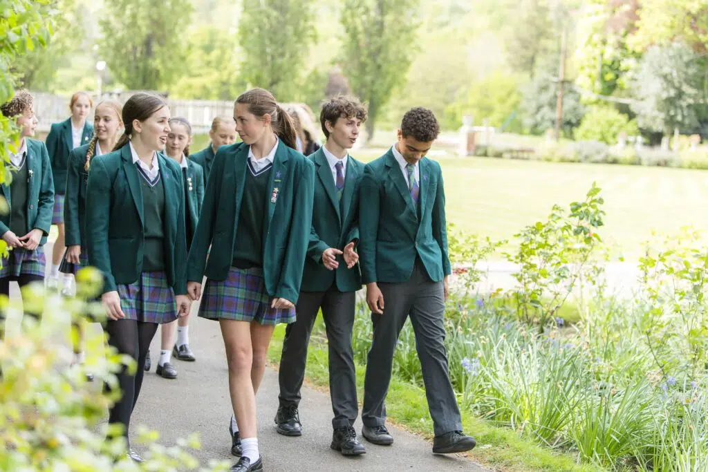 Group of students in green blazers walking along a park path outdoors.