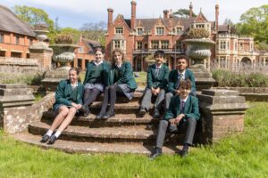 Group of students in green blazers and plaid skirts/pants sit on brick steps in a historic campus courtyard.