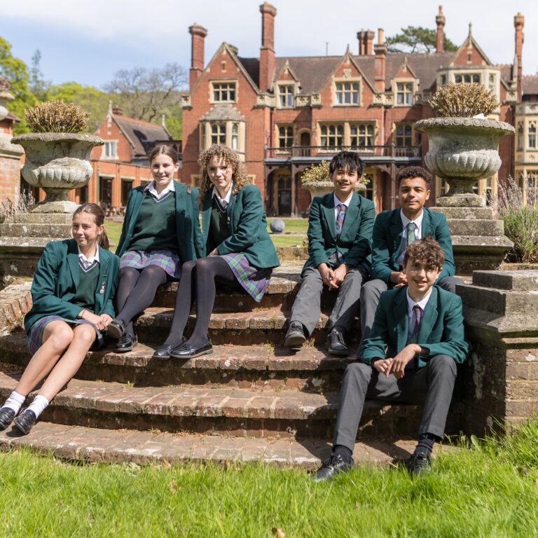 Group of students in green blazers and plaid skirts/pants sit on brick steps in a historic campus courtyard.
