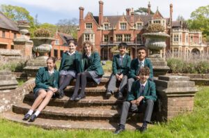Group of students in green blazers and plaid skirts/pants sit on brick steps in a historic campus courtyard.