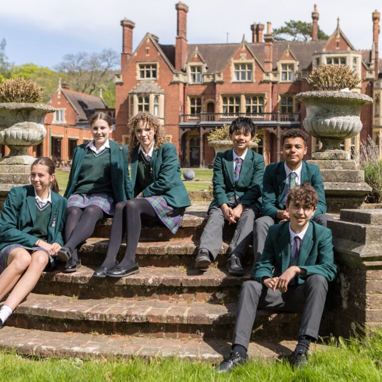 Group of students in green blazers and plaid skirts/pants sit on brick steps in a historic campus courtyard.
