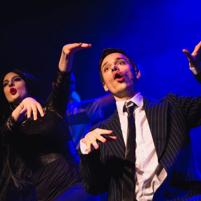 Two performers on stage under blue lighting; man in pinstripe suit singing and gesturing with raised hands, woman beside him in black dress.