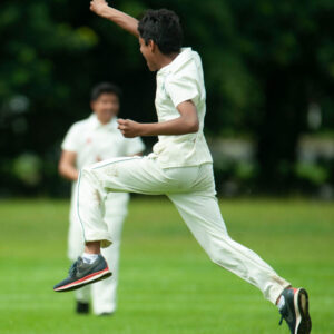 A man in a white cricket uniform jumps mid‑stride on a grassy field, with another player blurred in the background.