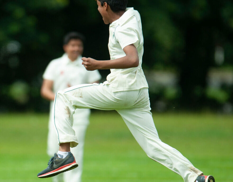 A man in a white cricket uniform jumps mid‑stride on a grassy field, with another player blurred in the background.