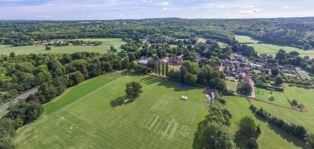 Aerial view of a green landscape with sports fields, trees, and a small village along a winding road under a blue sky.