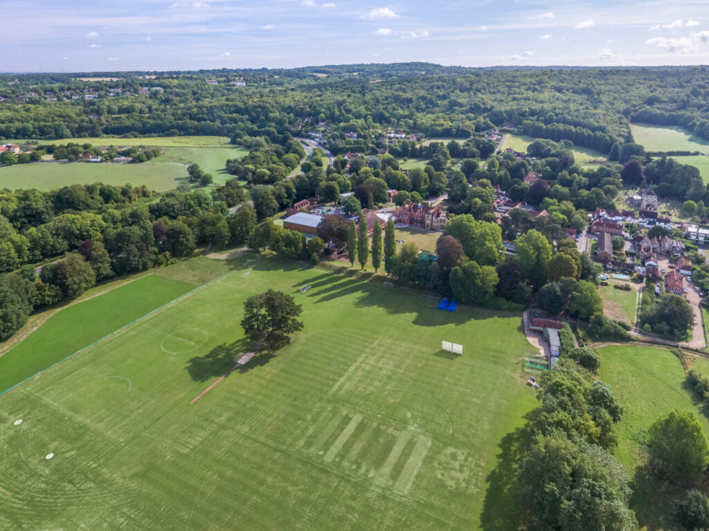 Aerial view of a green landscape with sports fields, trees, and a small village along a winding road under a blue sky.