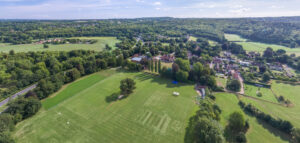 Aerial view of a green landscape with sports fields, trees, and a small village along a winding road under a blue sky.