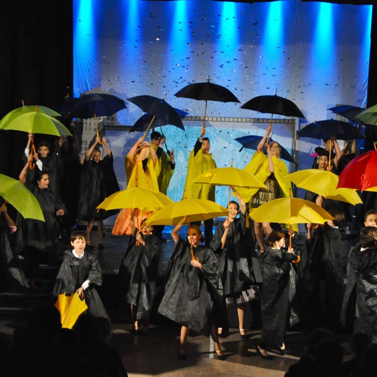 Group of children on a stage holding colorful umbrellas (red, yellow, green, blue) during a performance under blue stage lights.