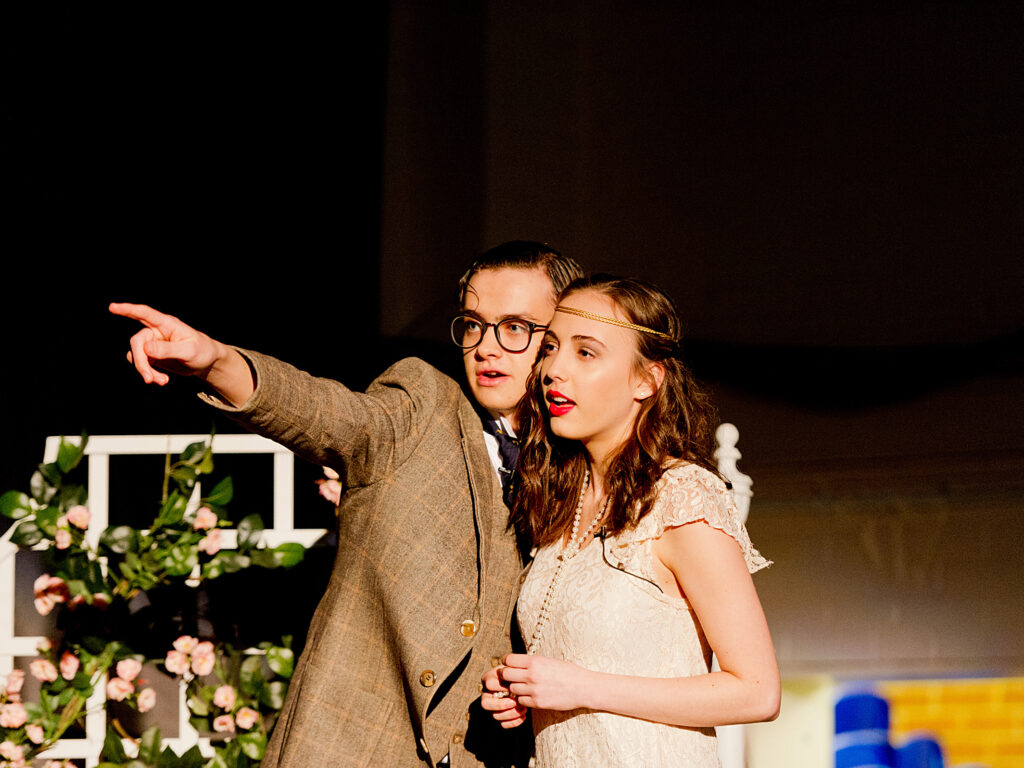 Man in a brown blazer and glasses points off to the left while standing with a woman in a cream lace dress; pink flowers and white decor visible in front (stage scene).