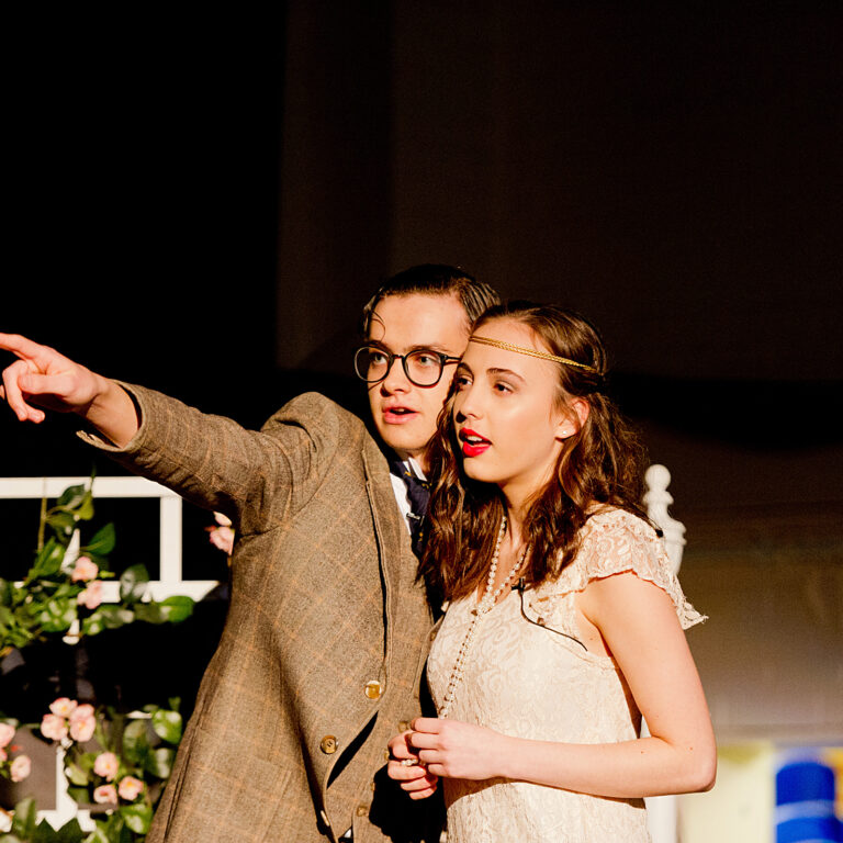 Man in a brown blazer and glasses points off to the left while standing with a woman in a cream lace dress; pink flowers and white decor visible in front (stage scene).