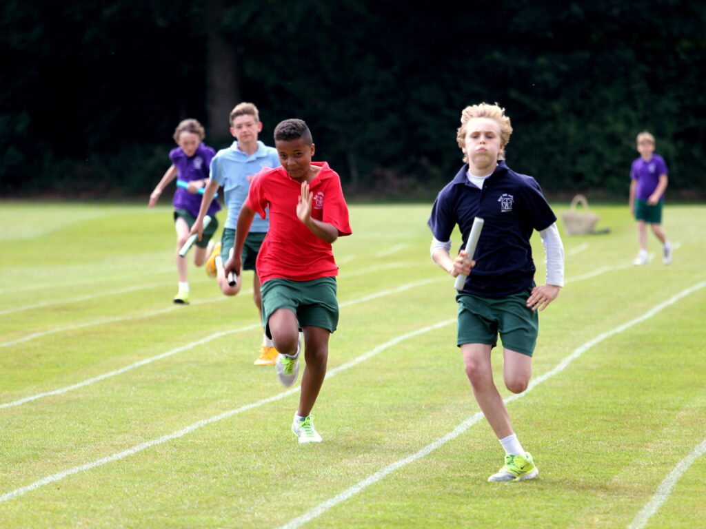 Children sprinting on a grassy field during a relay race, holding batons with a line of runners behind them
