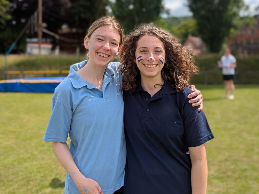 Two young women smiling and posing together on a sunny field, with blue face paint on their cheeks and arms around each other.