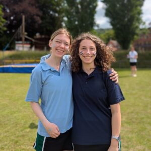 Two young women smiling and posing together on a sunny field, with blue face paint on their cheeks and arms around each other.