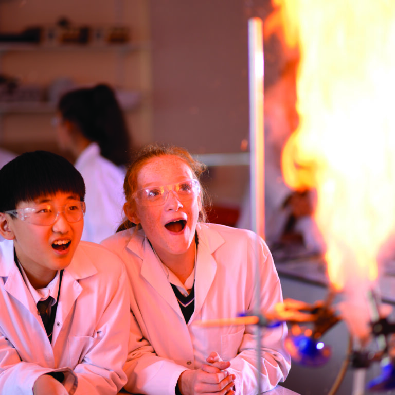 Two students in lab coats and safety goggles watch a fiery experiment in a science lab.