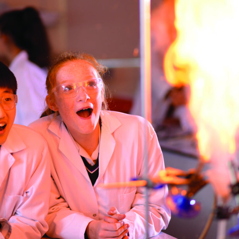 Two students in lab coats and safety goggles watch a fiery experiment in a science lab.