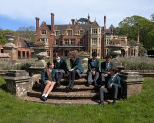 Group of students in green blazers seated on circular stone steps in front of a red brick manor and manicured garden.