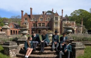 Group of students in green blazers seated on circular stone steps in front of a red brick manor and manicured garden.
