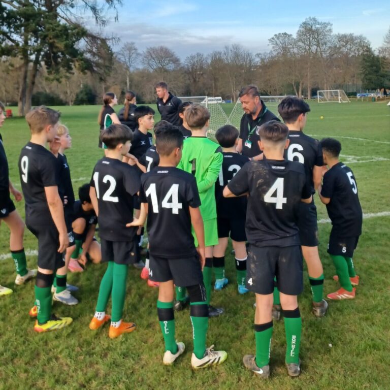 Youth soccer team huddled in a circle on a grassy field, players in black uniforms with green socks and numbers visible on backs.