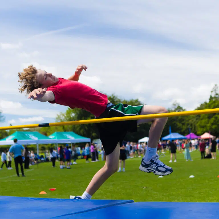 Athlete in a red shirt clears a yellow high jump bar outdoors, with spectators and tents in the background.”, “Young jumper in red shirt mid-air over a high jump bar at an outdoor track and field event.”, “Rowdy crowd watching a high jump attempt at a sunny outdoor sports event.”, “Athlete leaping over a high jump bar on a grassy field, with people and tents behind.”