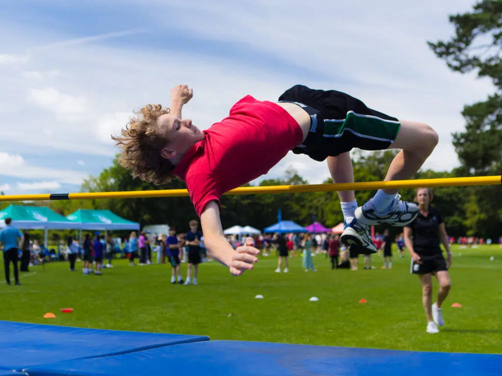 Boy in a red shirt clears a yellow high jump bar onto a blue mat at an outdoor field event, with spectators in the background on a sunny day.