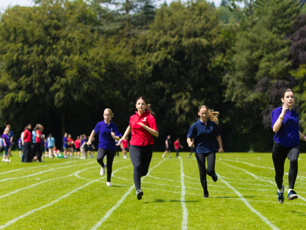 Group of students running on a grassy outdoor track during a race, with trees in the background.