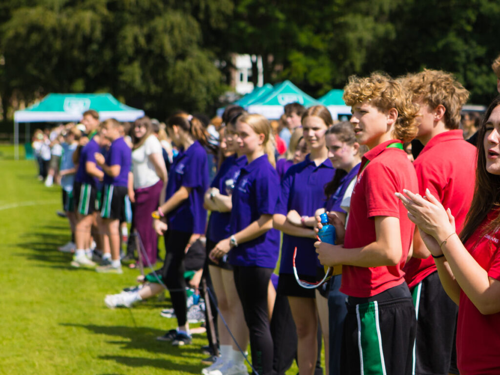 Line of young athletes in red and purple shirts standing in a row on a grassy field during an outdoor sports event, holding bows.