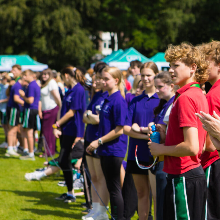 Line of young athletes in red and purple shirts standing in a row on a grassy field during an outdoor sports event, holding bows.