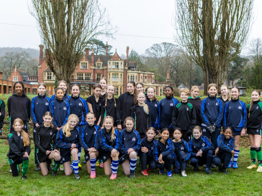 Group of girls in blue and black athletic uniforms posing for a team photo on a grassy field with a brick campus building in the background.