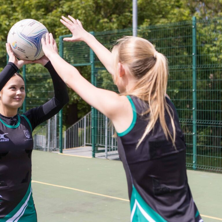 Two female players in black-green uniforms on an outdoor court reach to catch a white ball together mid-play. World Netball logo visible on ball.