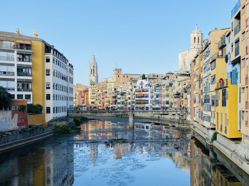 Colorful apartment buildings line a calm canal with reflections and a clear blue sky. Distant church towers rise above the urban skyline.