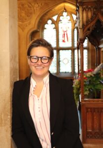 Smiling person with short hair and glasses wearing a blazer inside a church with stained-glass windows behind them