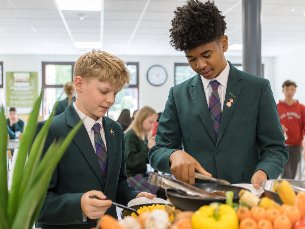 Two students in green blazers serve themselves at a school cafeteria buffet with colorful fruits and vegetables in front of them.
