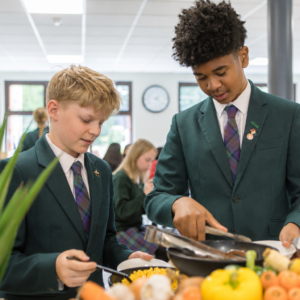 Two students in green blazers serve themselves at a school cafeteria buffet with colorful fruits and vegetables in front of them.